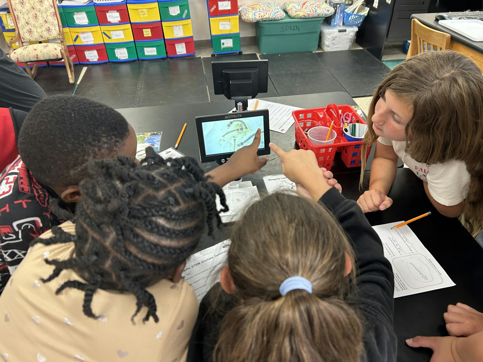 A group of children are engaged and pointing at a microscope, likely discussing the mosquito life cycle and mosquito larvae.
