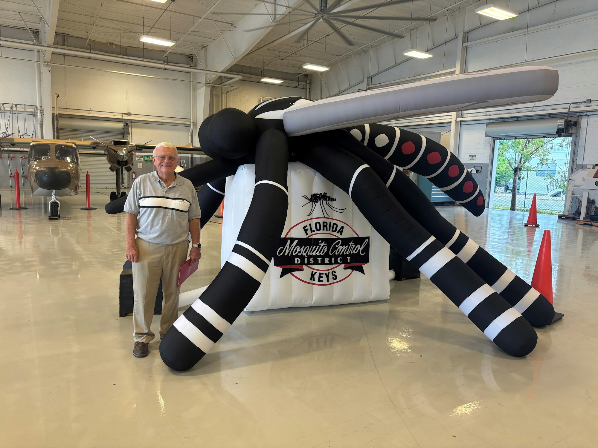 An inflatable mosquito model, a man posing beside it, and a helicopter in a hangar with a sign for Florida Mosquito Control.