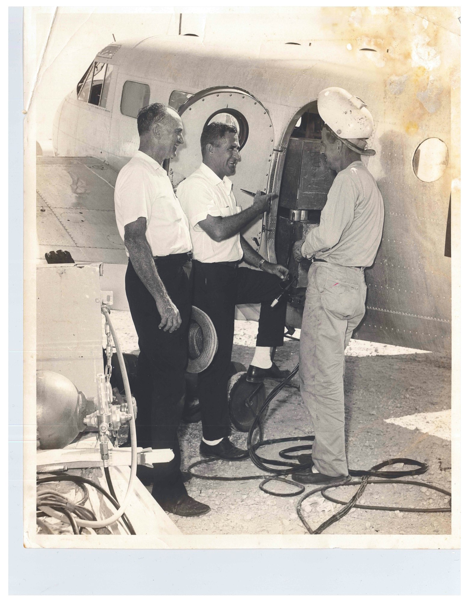 Three men examining equipment at an airplane's door; two wear uniforms, one in work attire with a helmet. Cables and devices visible.