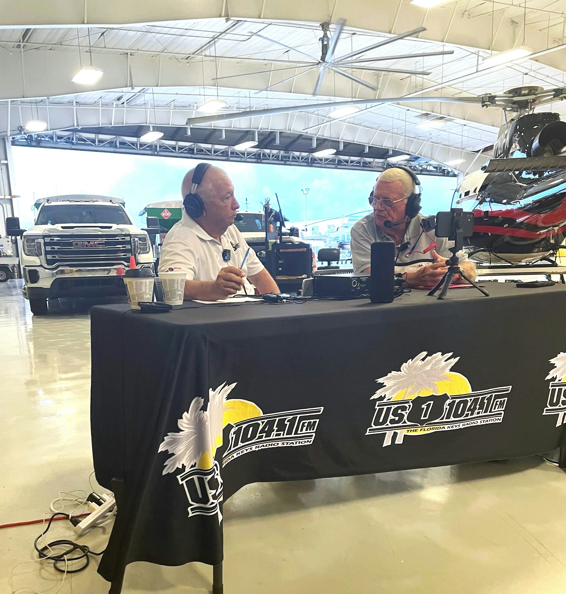 Two people with headphones in a hangar-like setting with a helicopter and vehicles in the background, likely a radio broadcast.