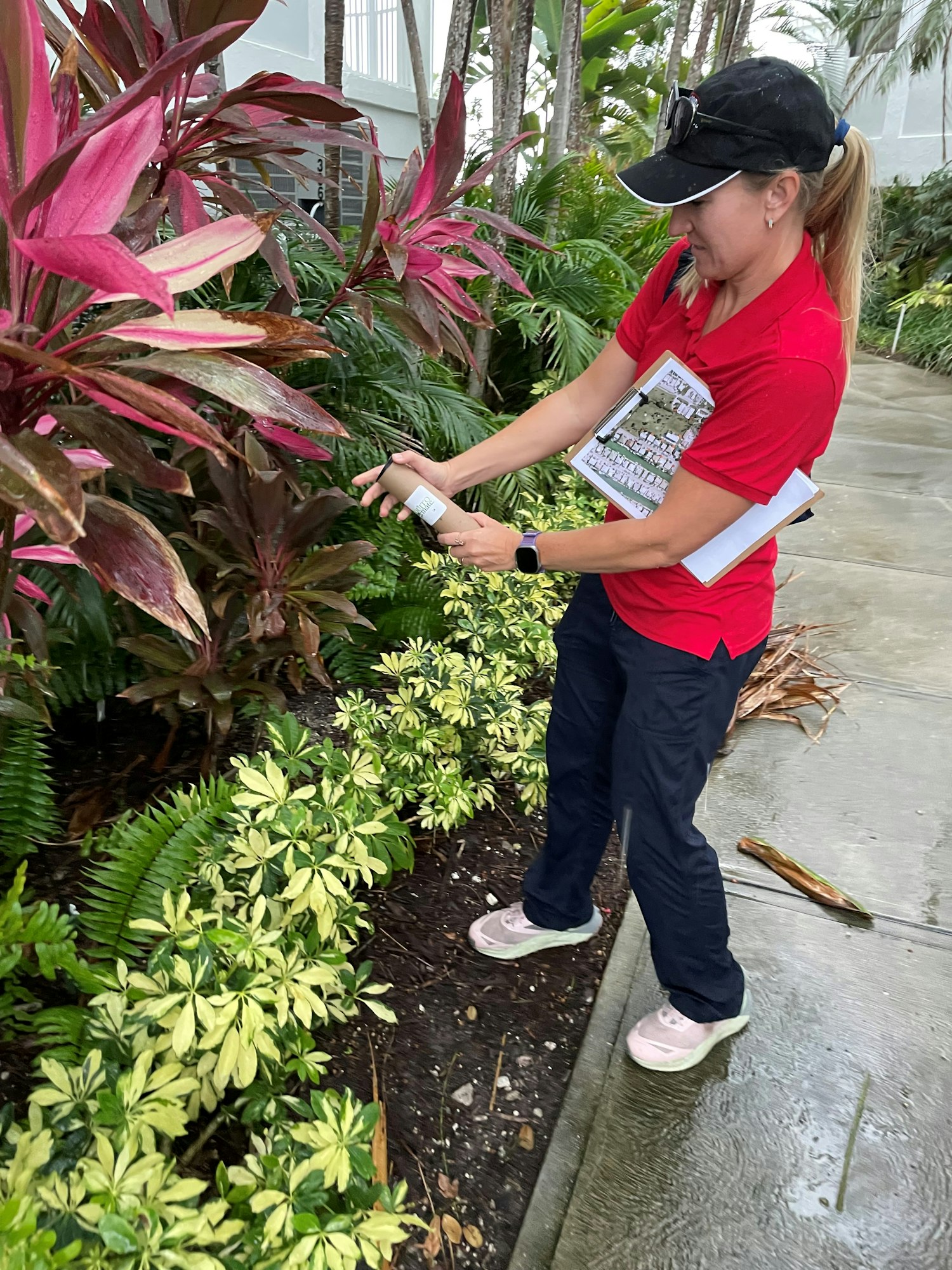 A person in a red shirt examines plants, holding a device and papers, surrounded by tropical foliage.