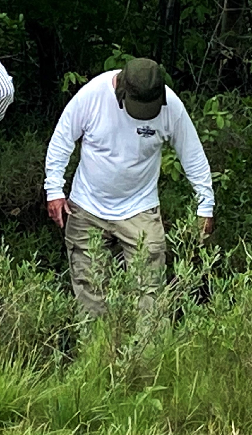 A person wearing a hat, long-sleeve shirt, and cargo pants stands in a grassy, wooded area.
