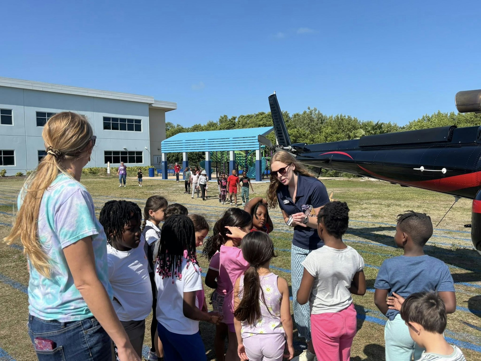 Kids are gathered outside near a helicopter, listening to an instructor, with a school building in the background.