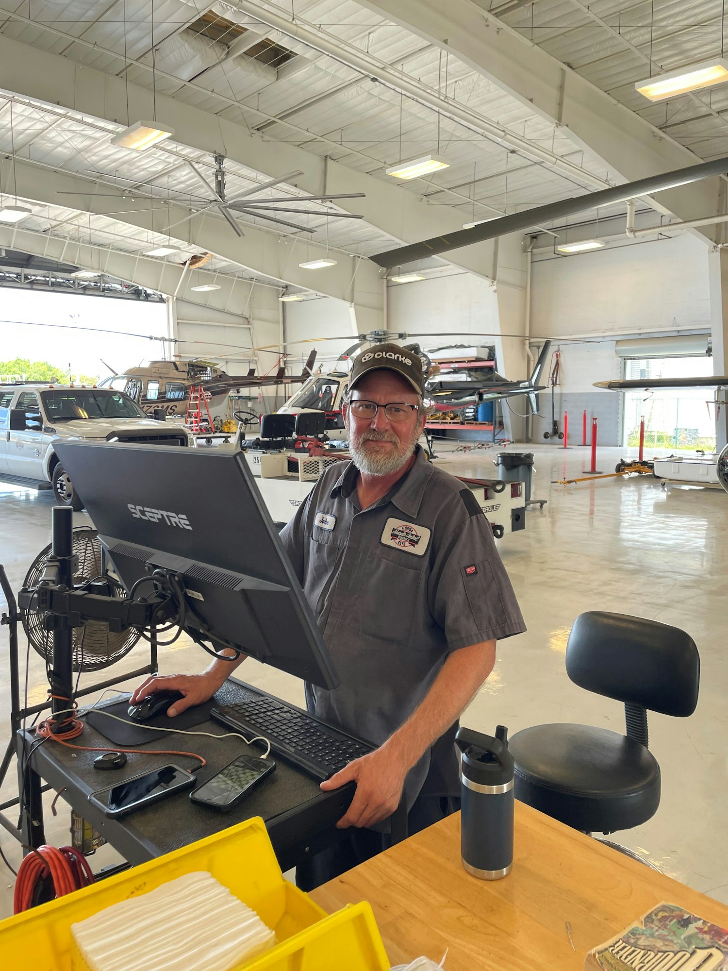 Man at a computer desk in a workshop; vehicles, equipment, and high ceiling in the background. (Image is rotated)