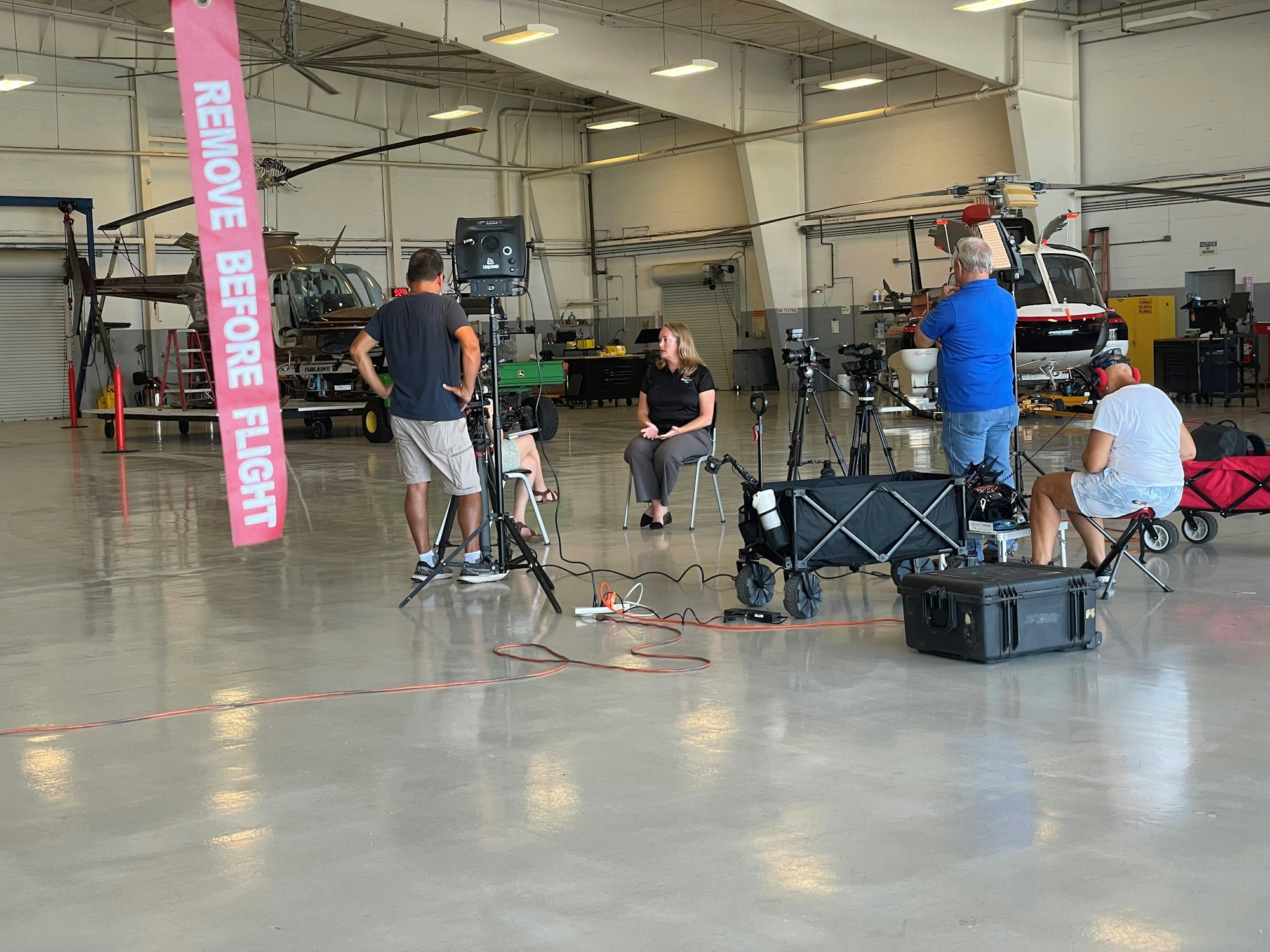 A video shoot is set up in a hangar with helicopters in the background. A pink "REMOVE BEFORE FLIGHT" tag hangs in the foreground.