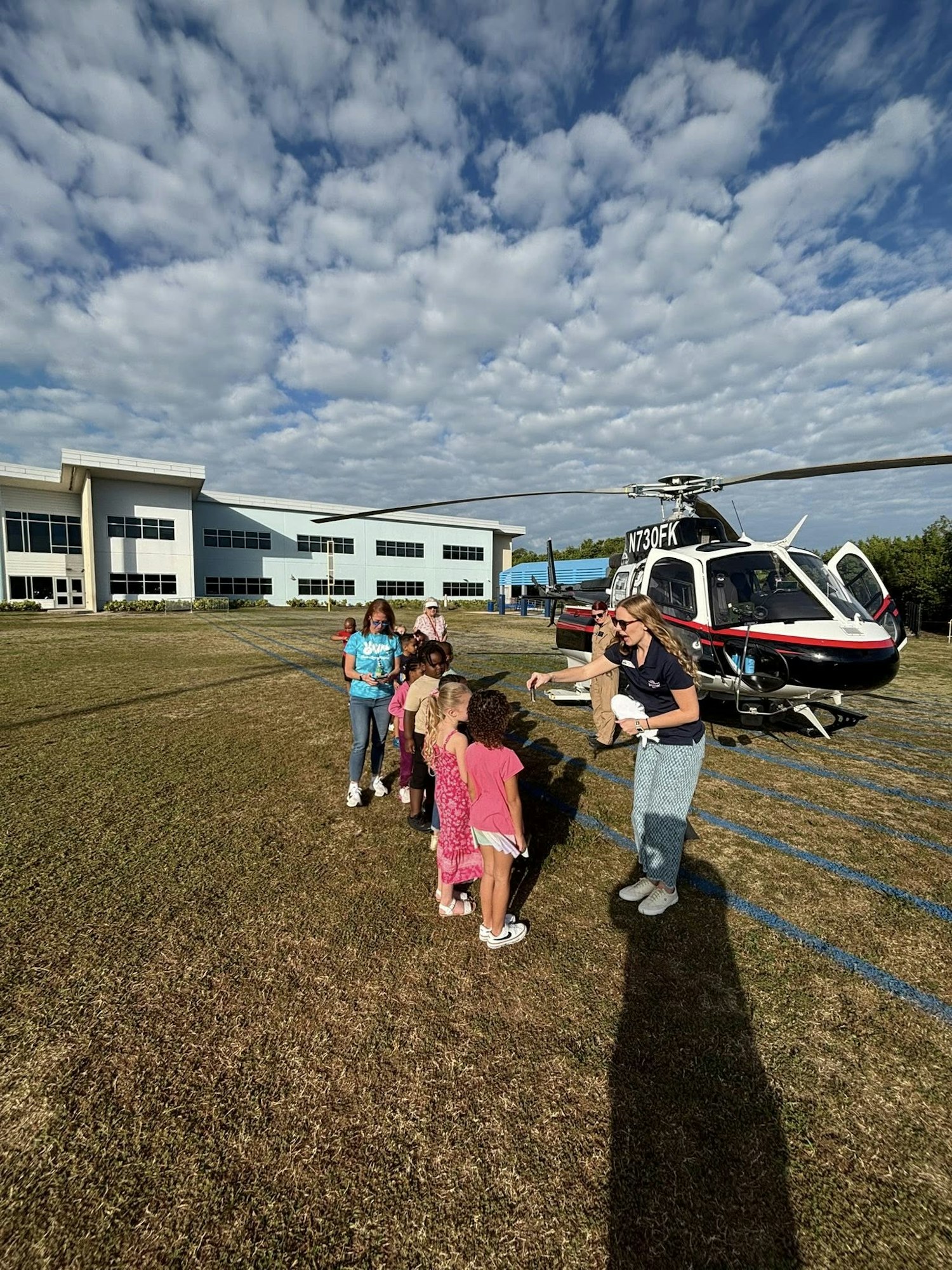 A helicopter on a grassy field with a group of children excitedly waiting nearby, and a person interacting with them.