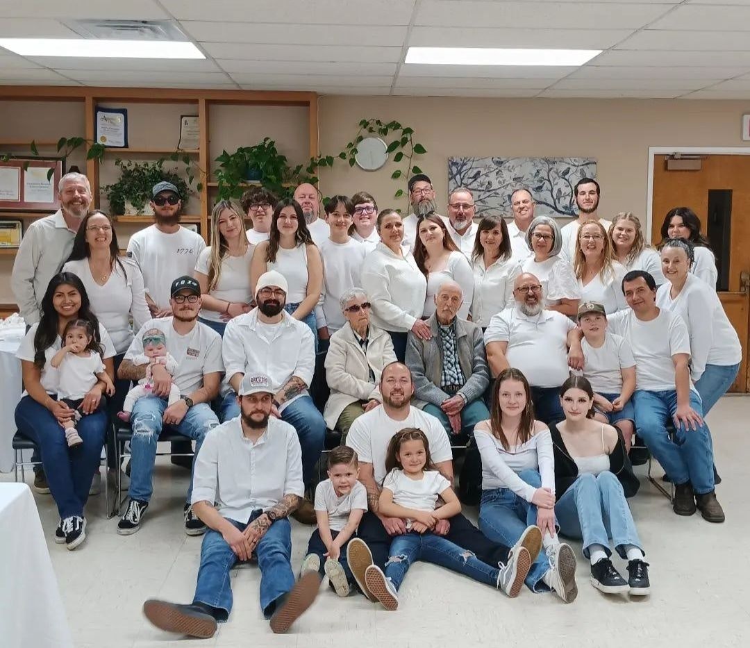 A large group of people posing for a photo, likely a family gathering, with most wearing white tops and smiling.