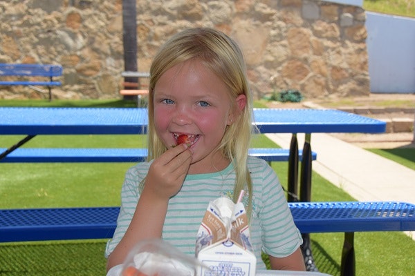 A blonde girl enjoys a snack at a picnic table, smiling and holding a piece of food, with a sunny outdoor background.