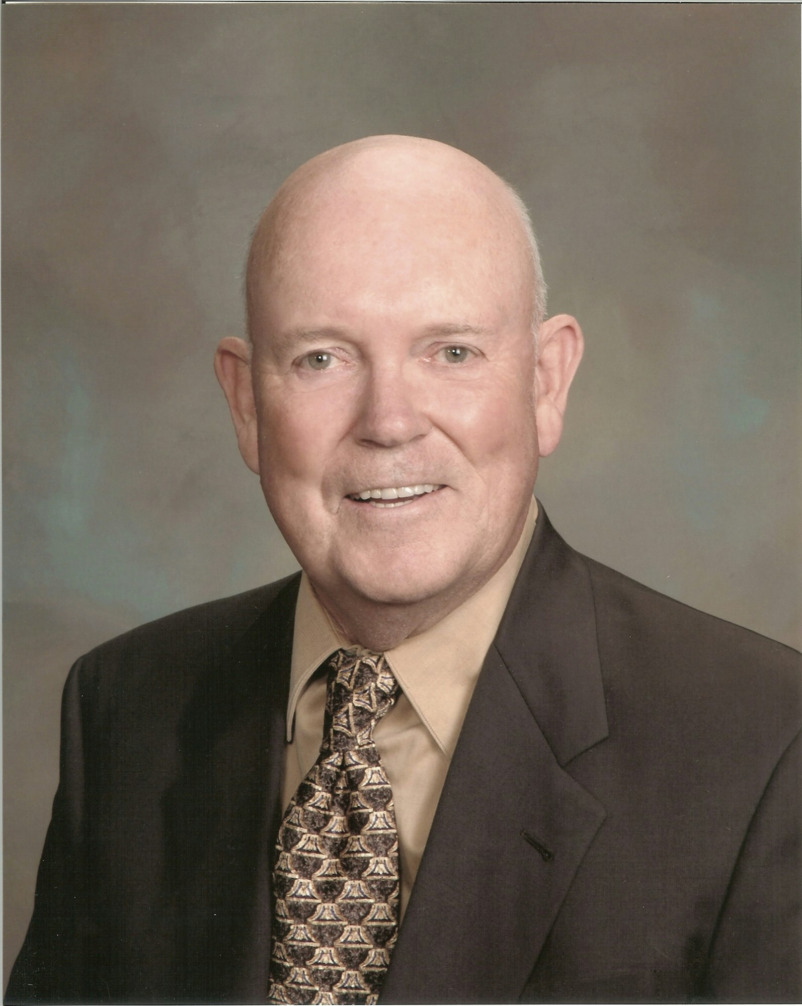 A smiling man in a suit with a patterned tie, posed against a neutral backdrop.