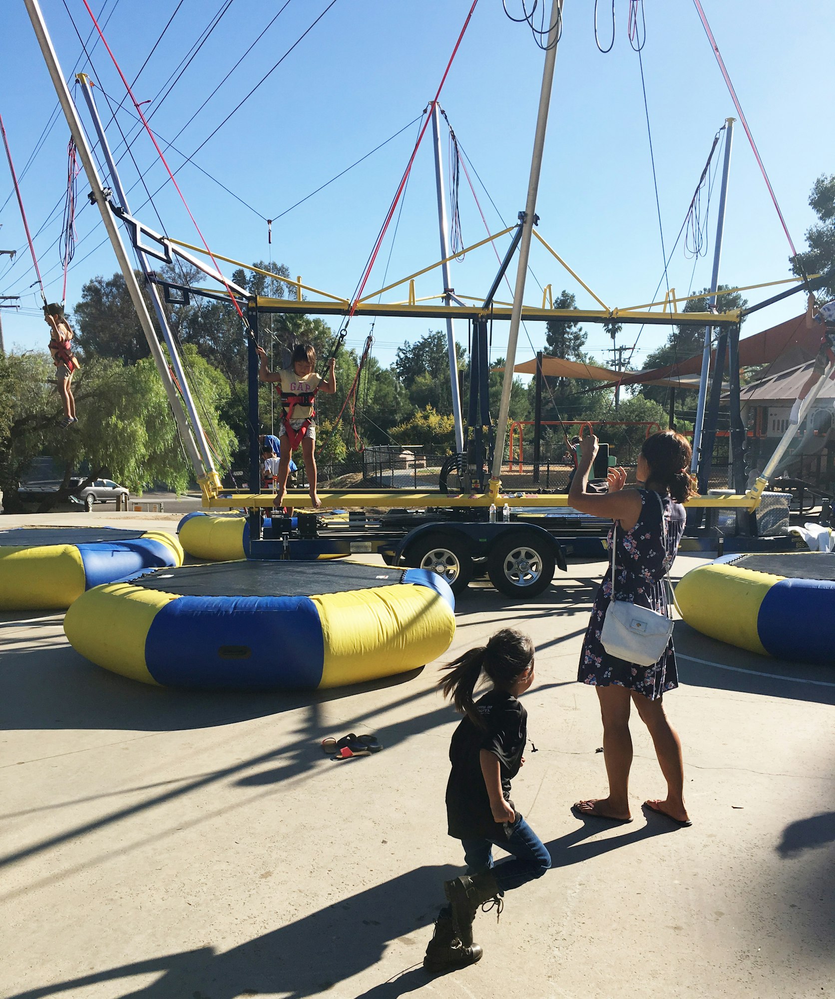 Children playing on a bungee trampoline, with one child jumping and an adult capturing the moment. Bright sunny day.