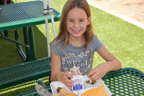 A young girl smiles while holding a milk carton and snacks on a tray, sitting at a picnic table on grass.