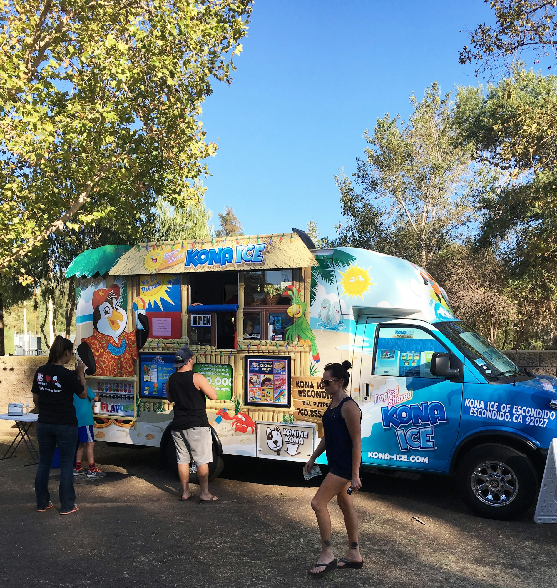 A colorful Kona Ice truck serving shaved ice with customers lined up, surrounded by trees and clear blue skies.