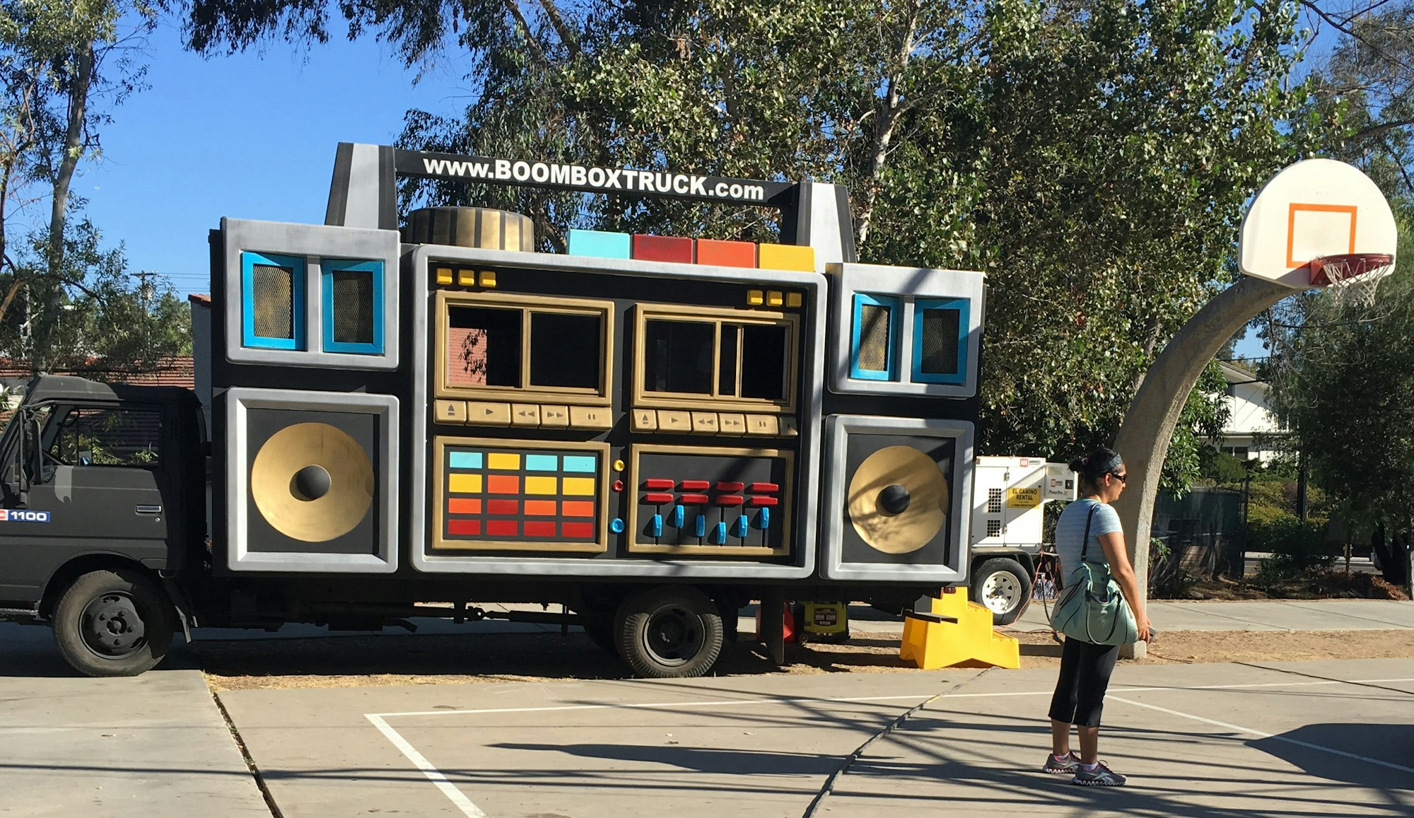 A large boombox truck is parked by a basketball court, with a woman standing nearby, enjoying the scene.