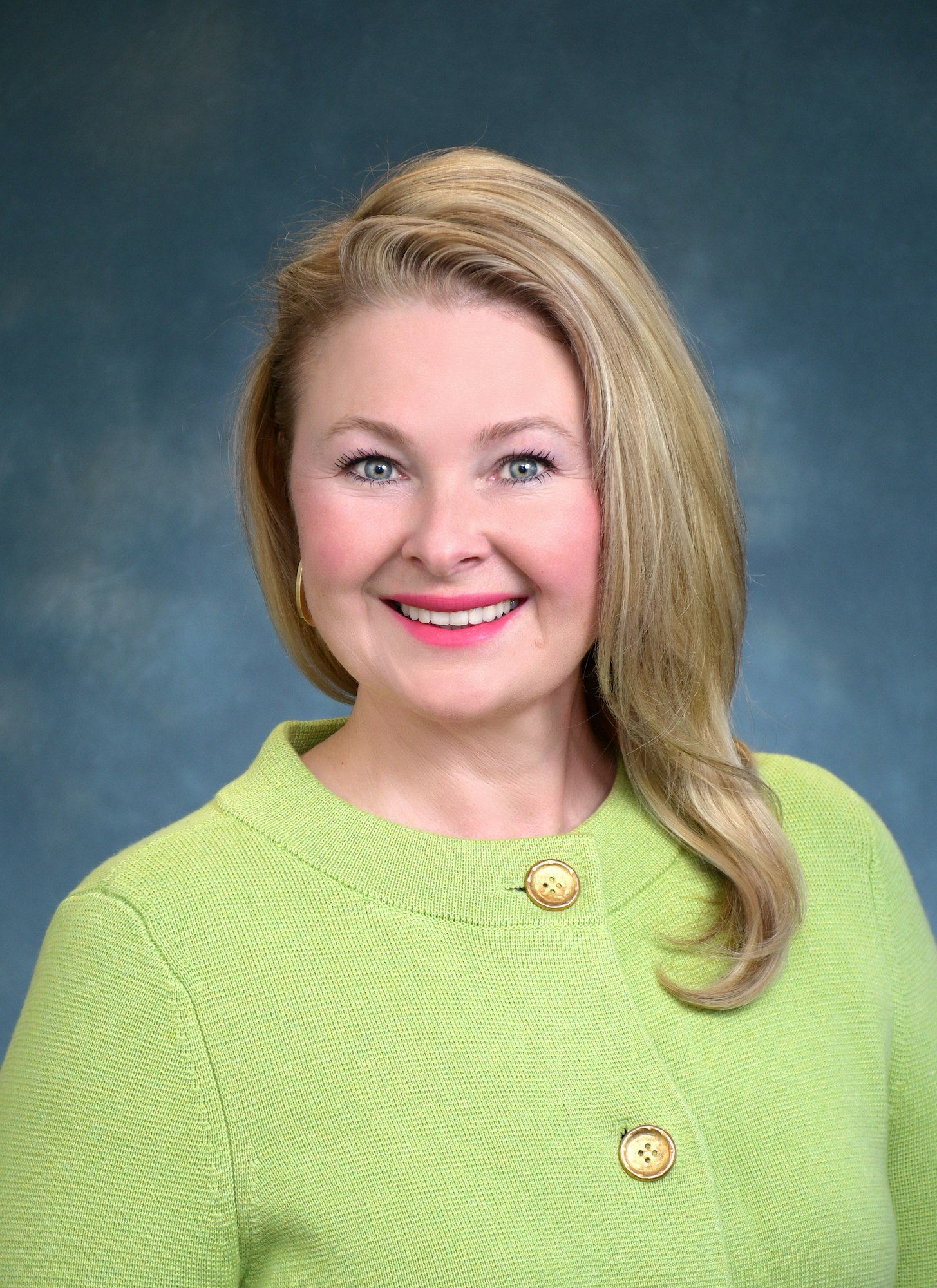 This is a picture of Olga, she's a smiling woman with blonde hair, wearing a green sweater with gold buttons, against a blue background.