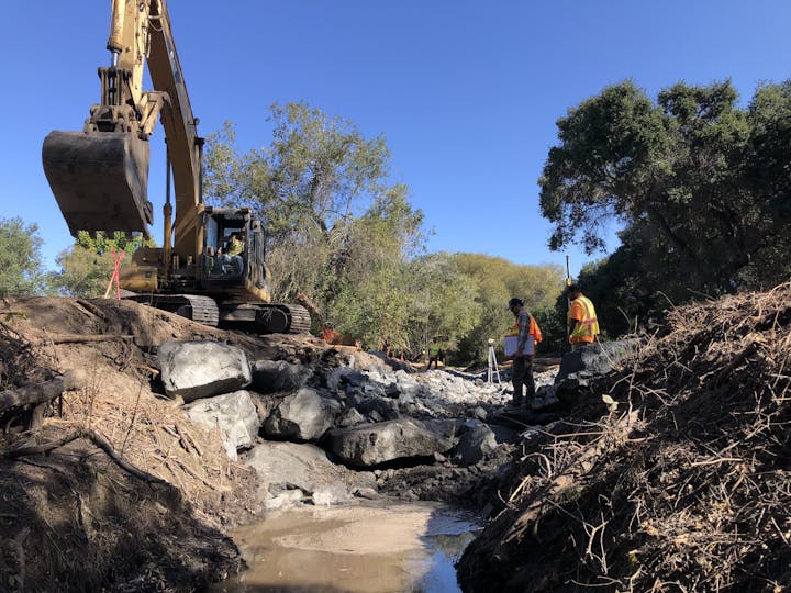 A construction scene with an excavator, two workers in safety vests, and rocky terrain surrounded by trees under a clear blue sky.