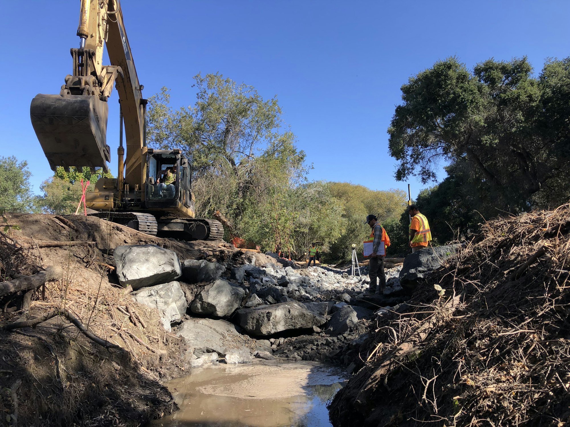 A construction scene with an excavator, two workers in safety vests, and rocky terrain surrounded by trees under a clear blue sky.