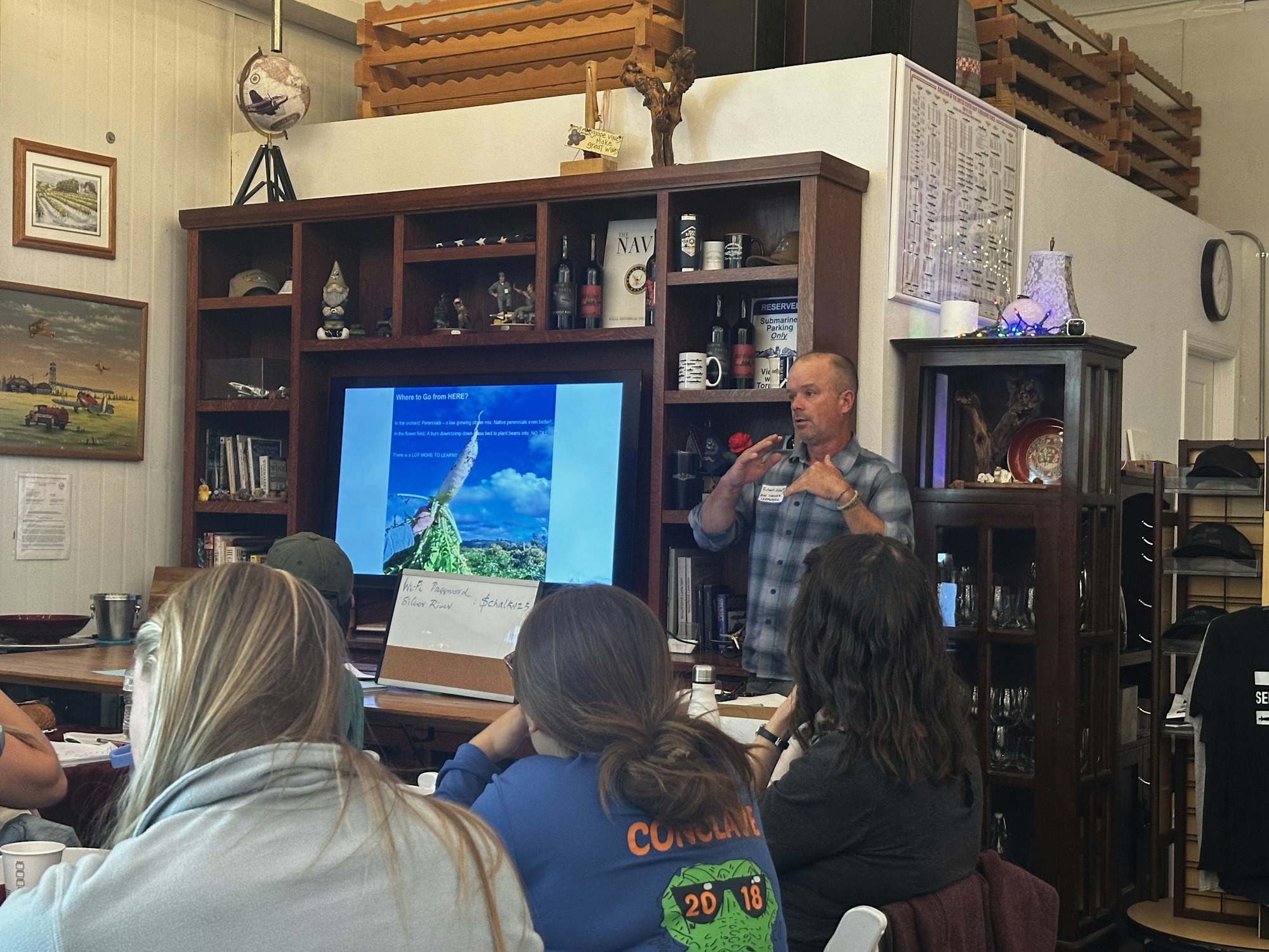 A speaker is giving a presentation to an audience in a cozy indoor setting, featuring visuals and a bulletin board.