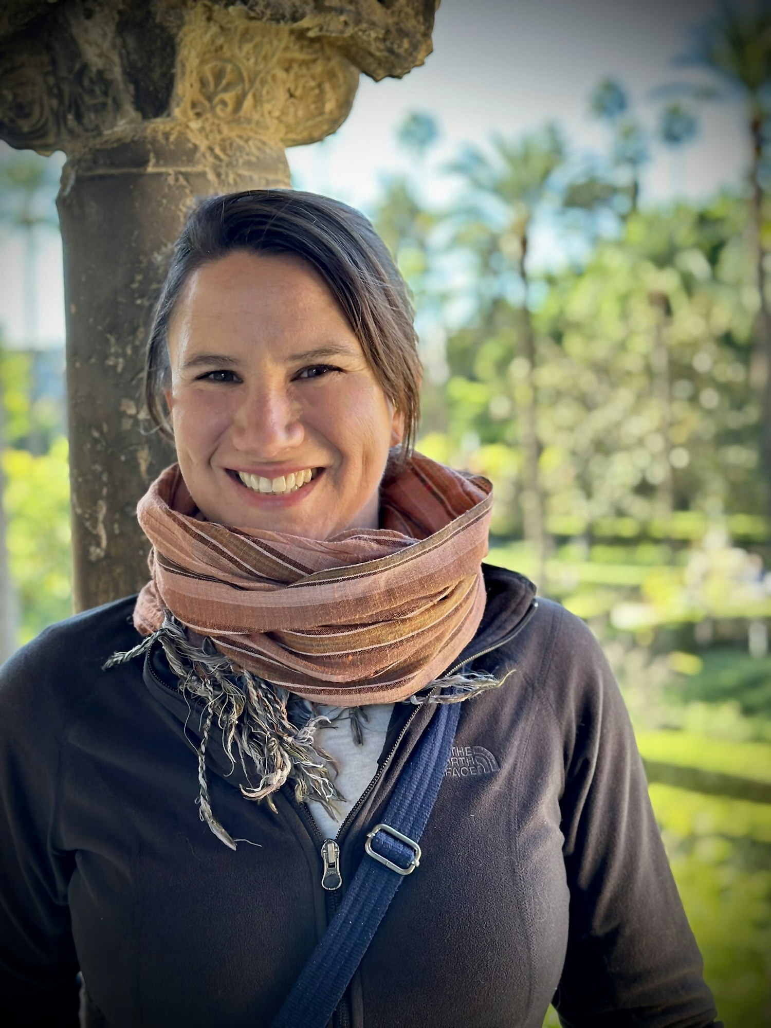 A person smiling, wearing a scarf, standing outdoors with trees and a decorative pillar behind them.