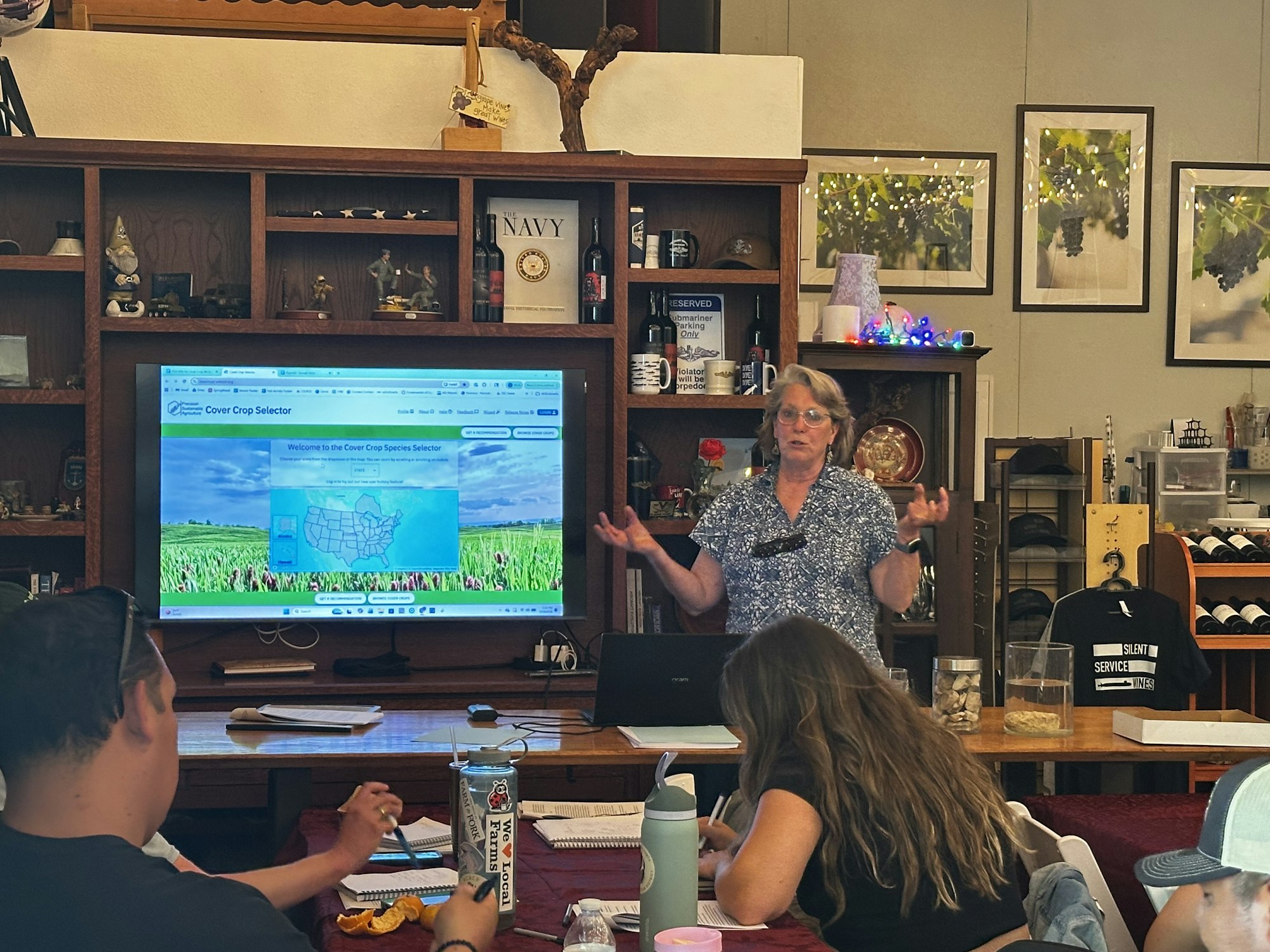 A woman is presenting about cover crops to an audience in a rustic setting, featuring a large screen with online content.