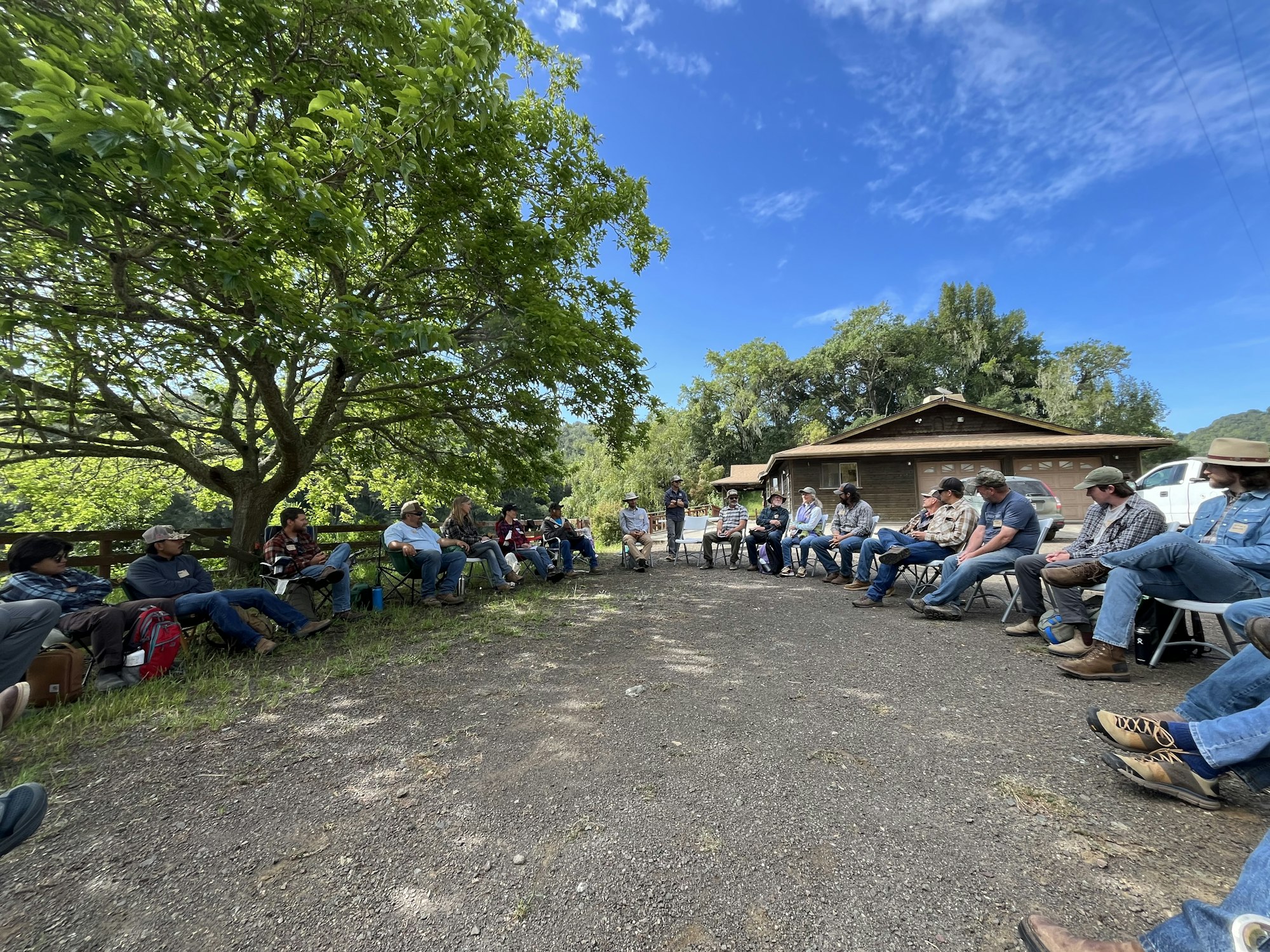 A group of people sitting in chairs outdoors near a house, engaged in a gathering under a large tree.
