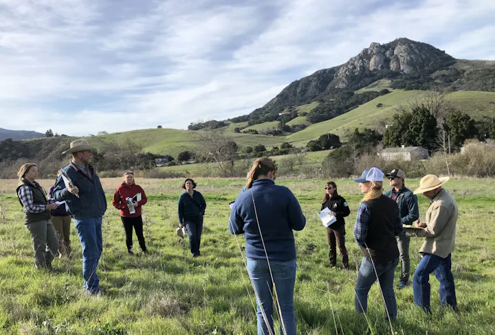 A group of people is gathered outdoors, discussing or learning in a scenic landscape with hills in the background.