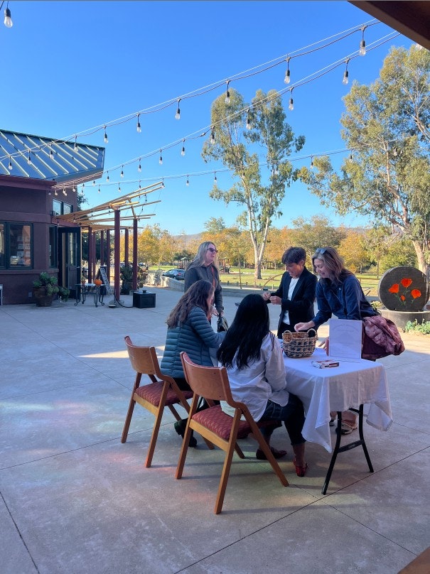 A group of people gathers outdoors at a table, engaging in conversation, with a bright blue sky and trees in the background.