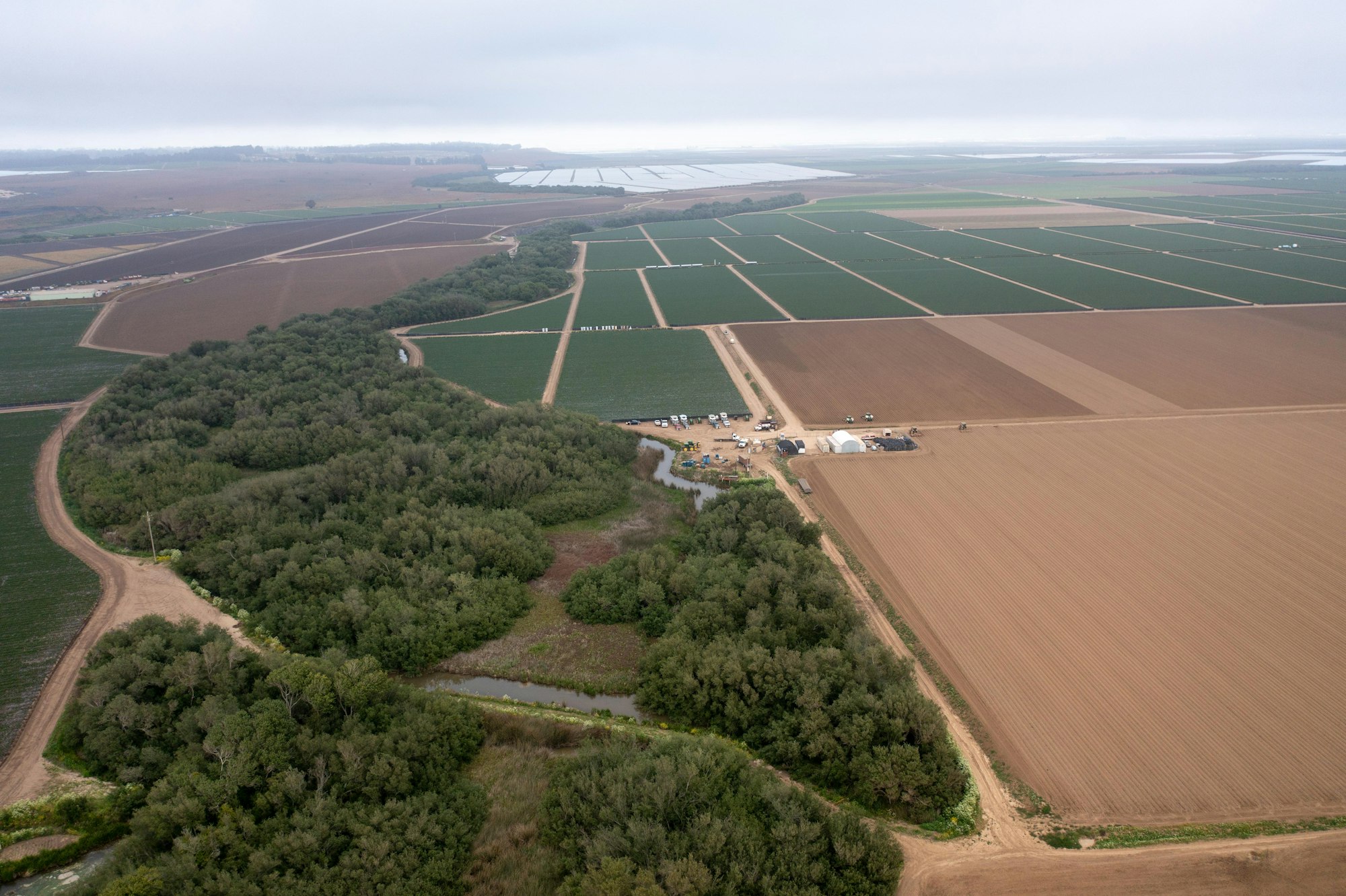 Aerial view of vast farmland with a mix of cultivated fields, forest, and a farm area with vehicles and structures.