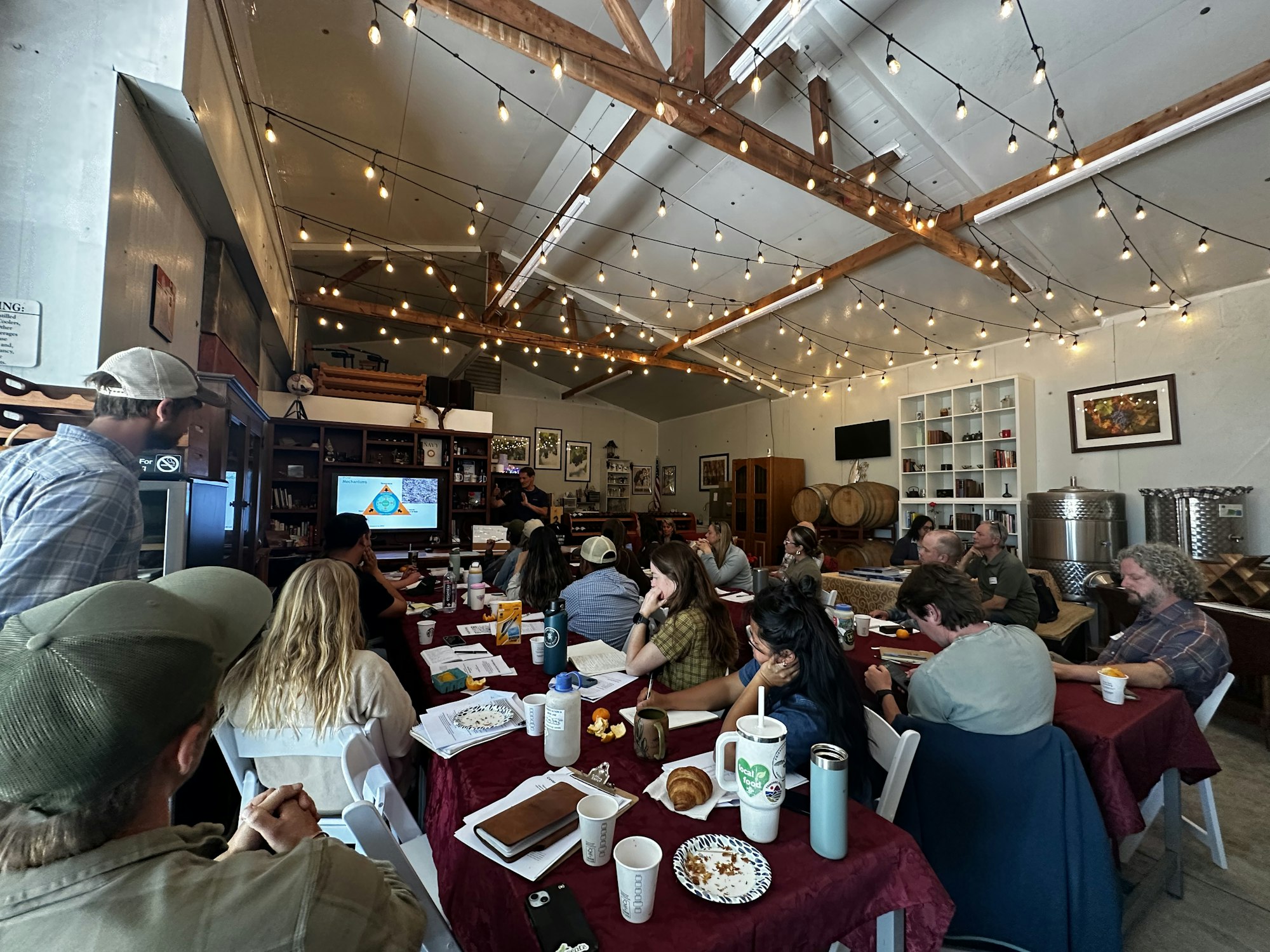 A group of people are seated in a cozy venue with a presentation, engaged in discussion over refreshments and notes.