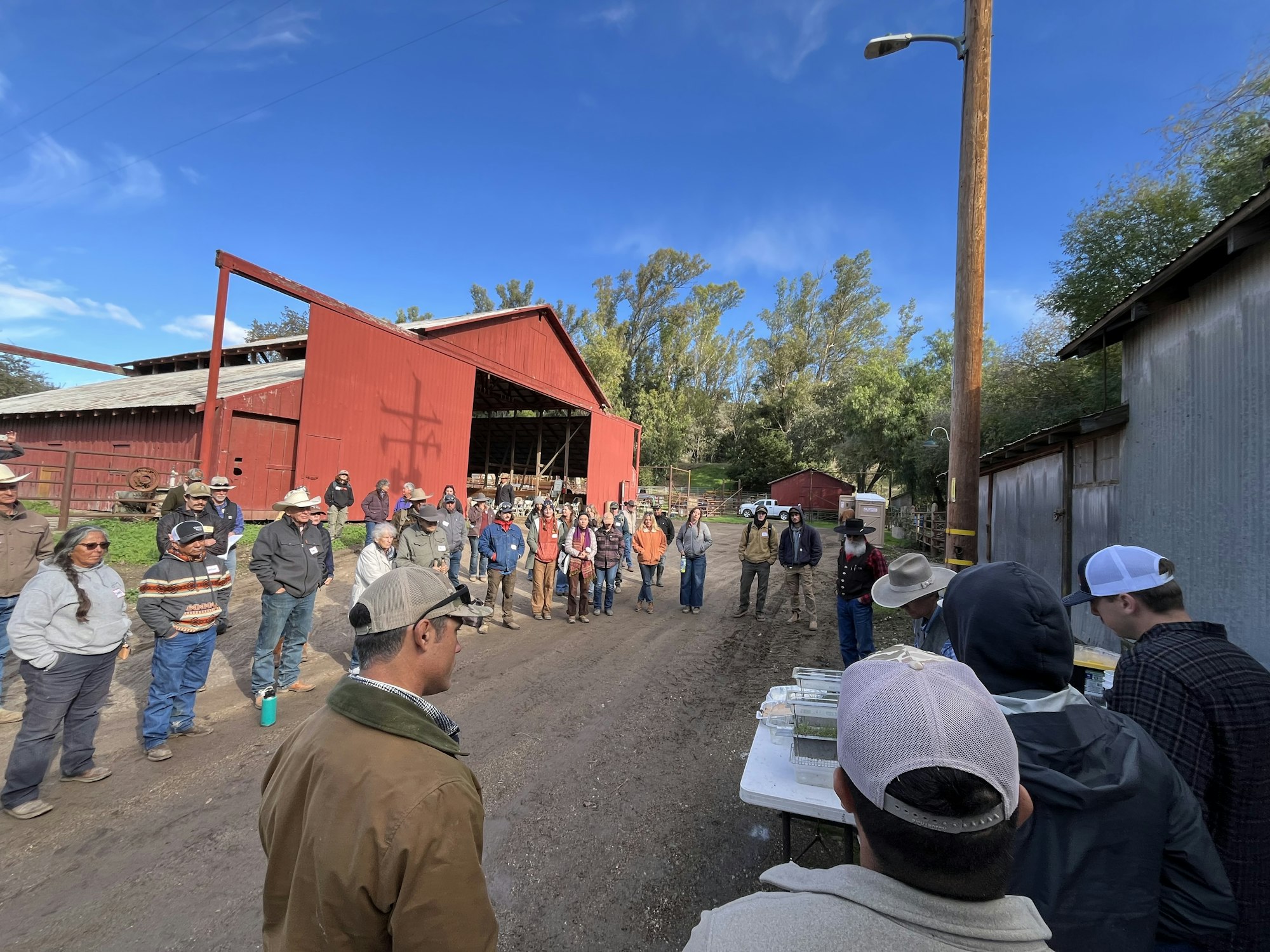 A group of people gathered outdoors near red barns, listening to a speaker, possibly for a workshop or training event.