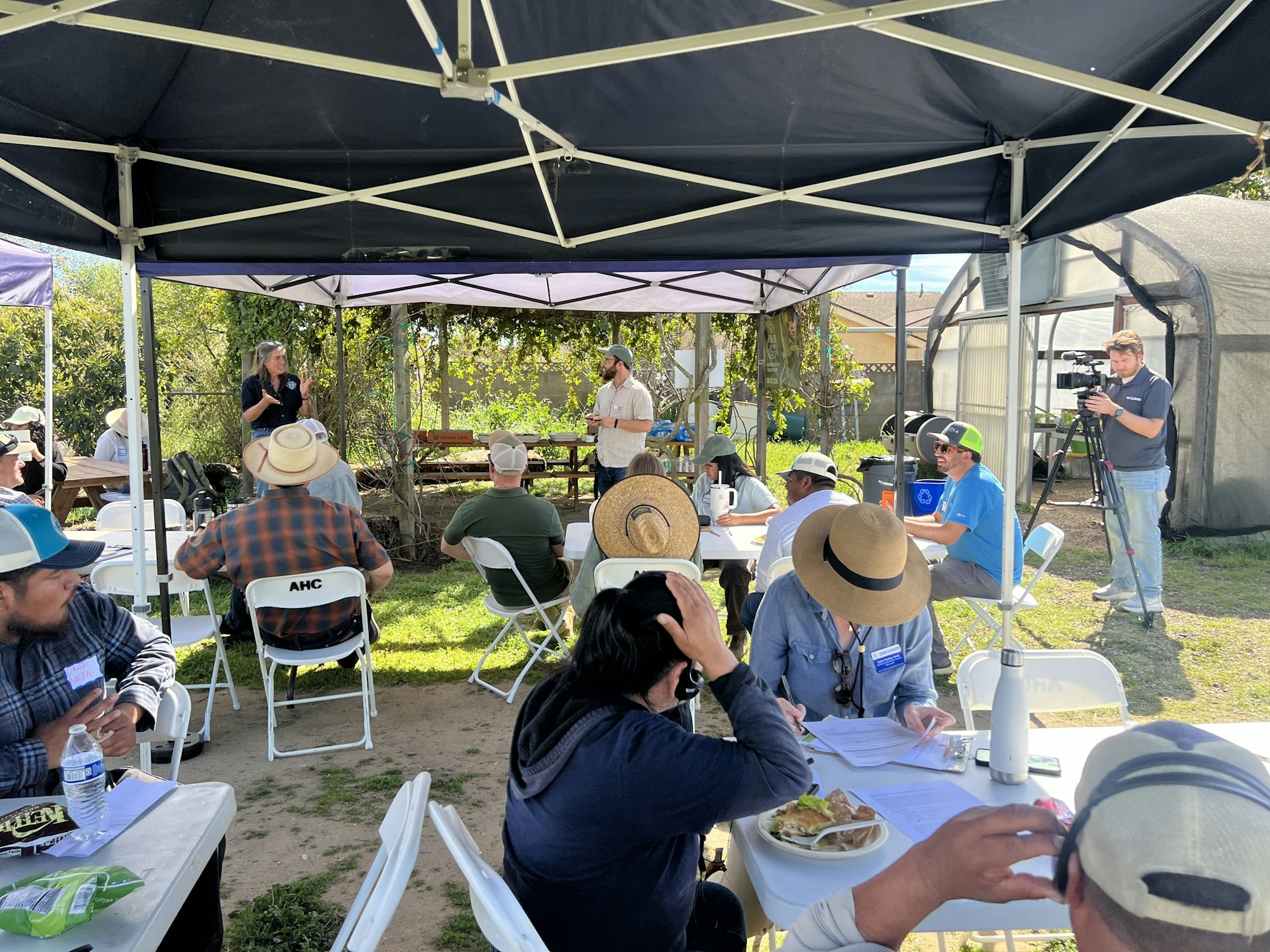 A gathering under a tent where individuals listen to a speaker, with people dining and a camera operator filming the event.