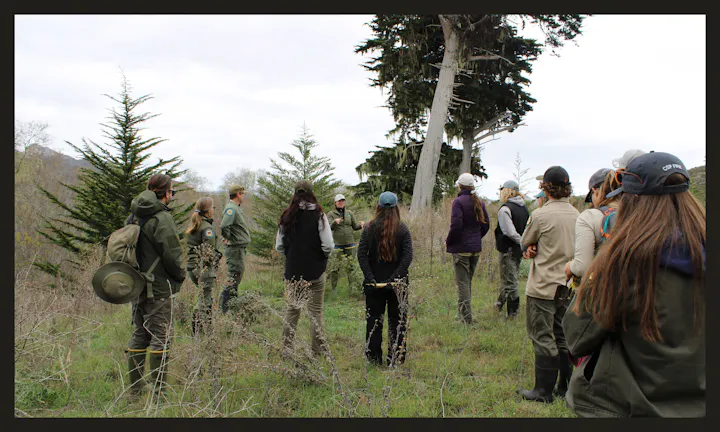 A group of people, in a nature setting, listen to a guide or instructor, surrounded by trees and greenery.