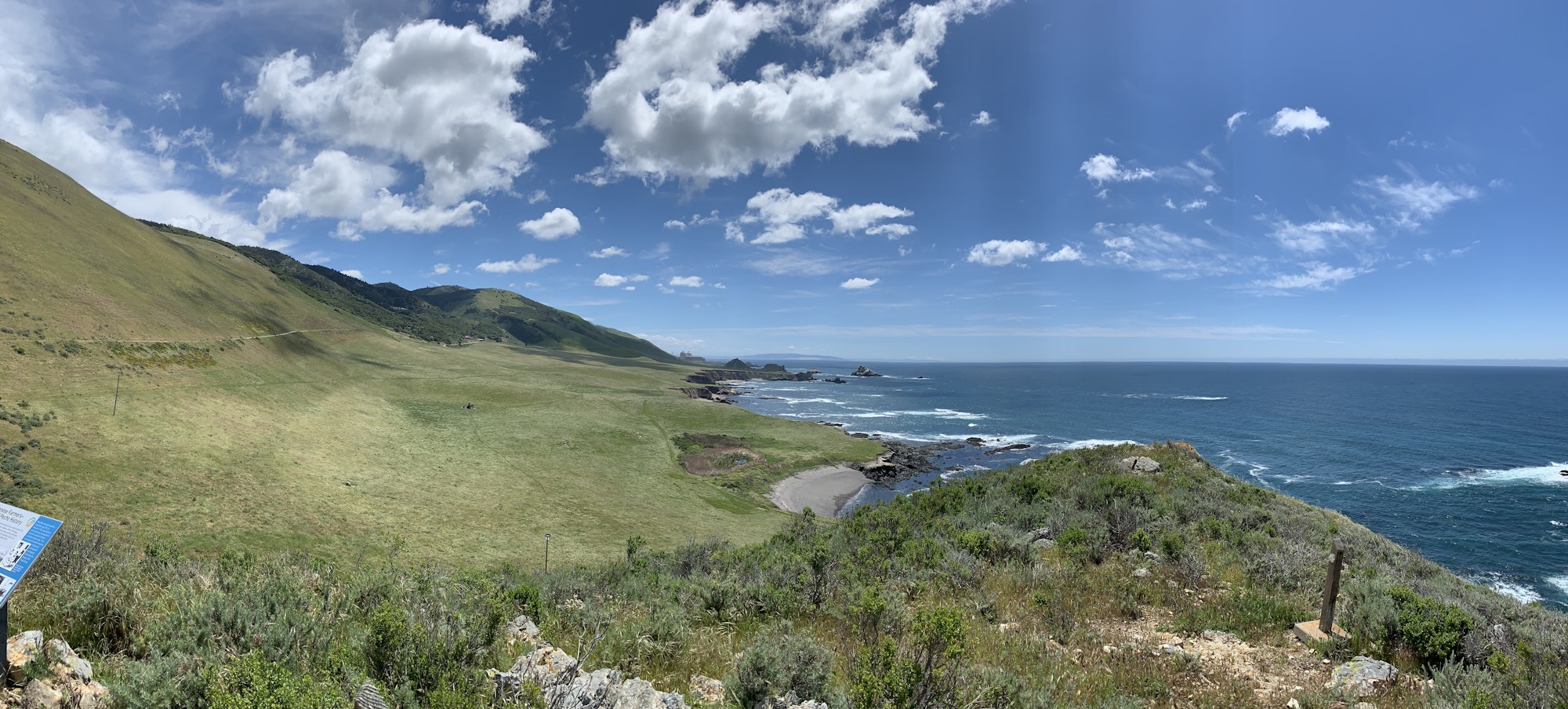 A panoramic view of coastal landscape with rolling green hills, rocky shores, ocean, and blue sky.