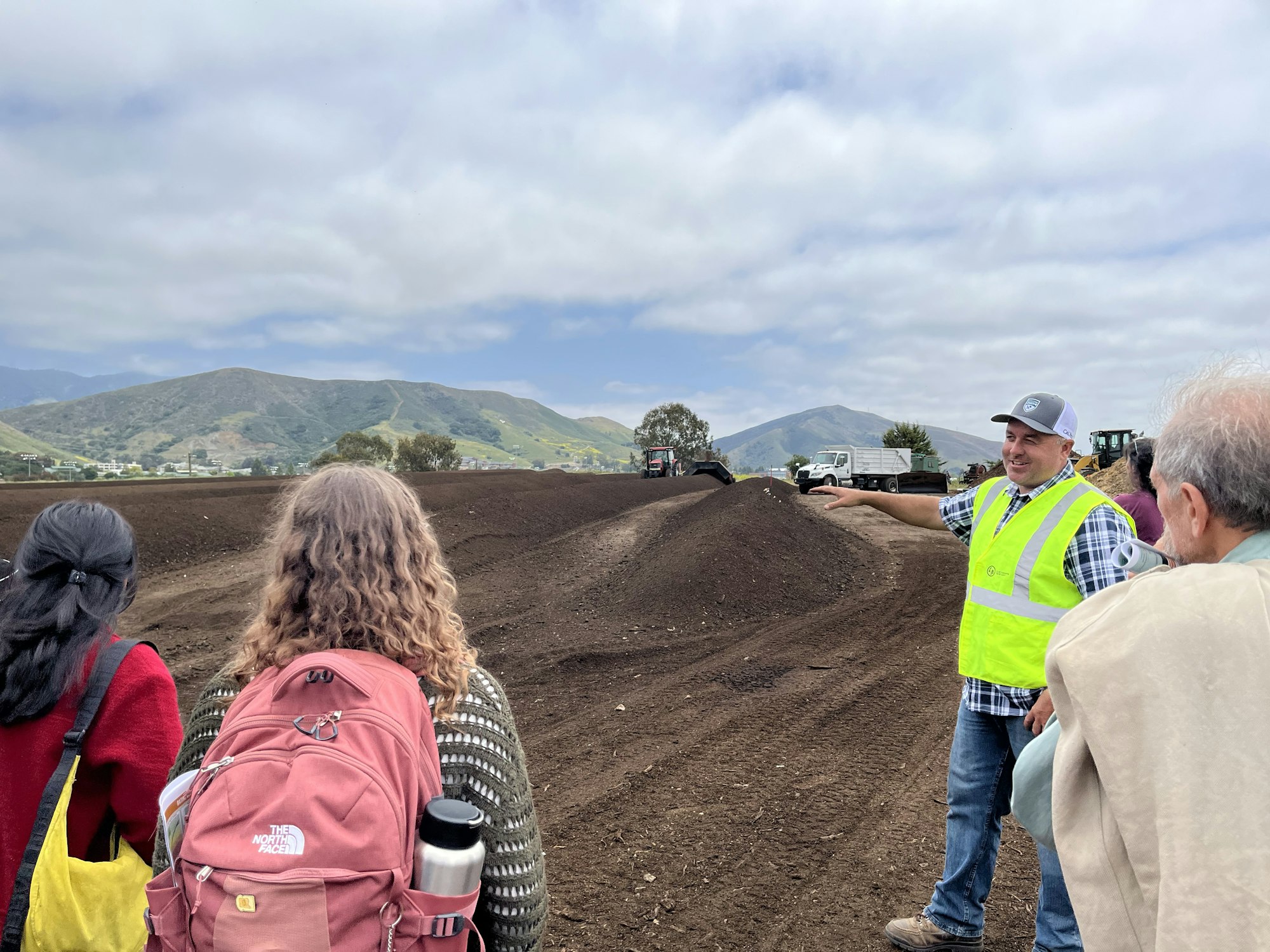 People outdoors near a dirt site, one in a safety vest. A truck and hilly landscape in the background.