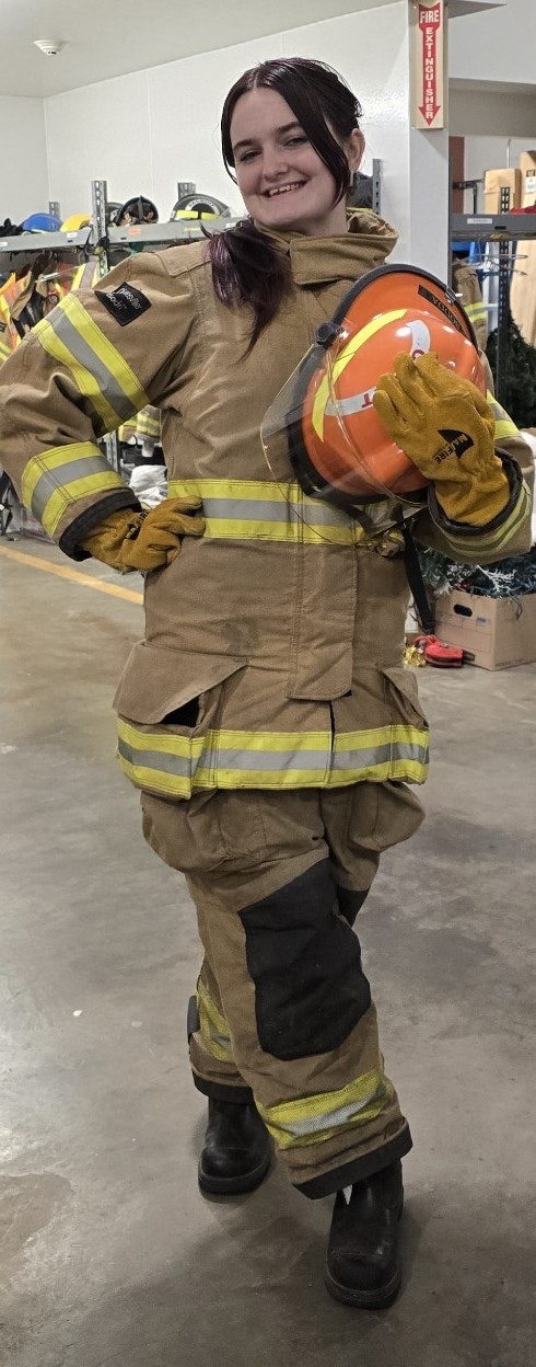 A person in a firefighting suit holding a helmet, smiling, in a storage room with shelves and various equipment.