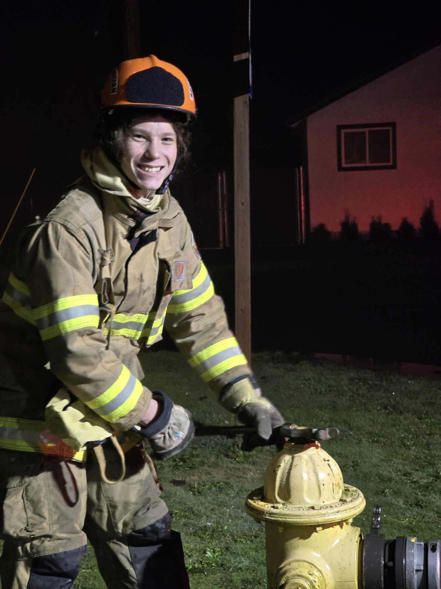 A firefighter in gear smiling while opening a yellow fire hydrant at night.