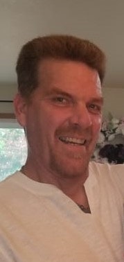 A smiling man with short hair in a white T-shirt, indoors.