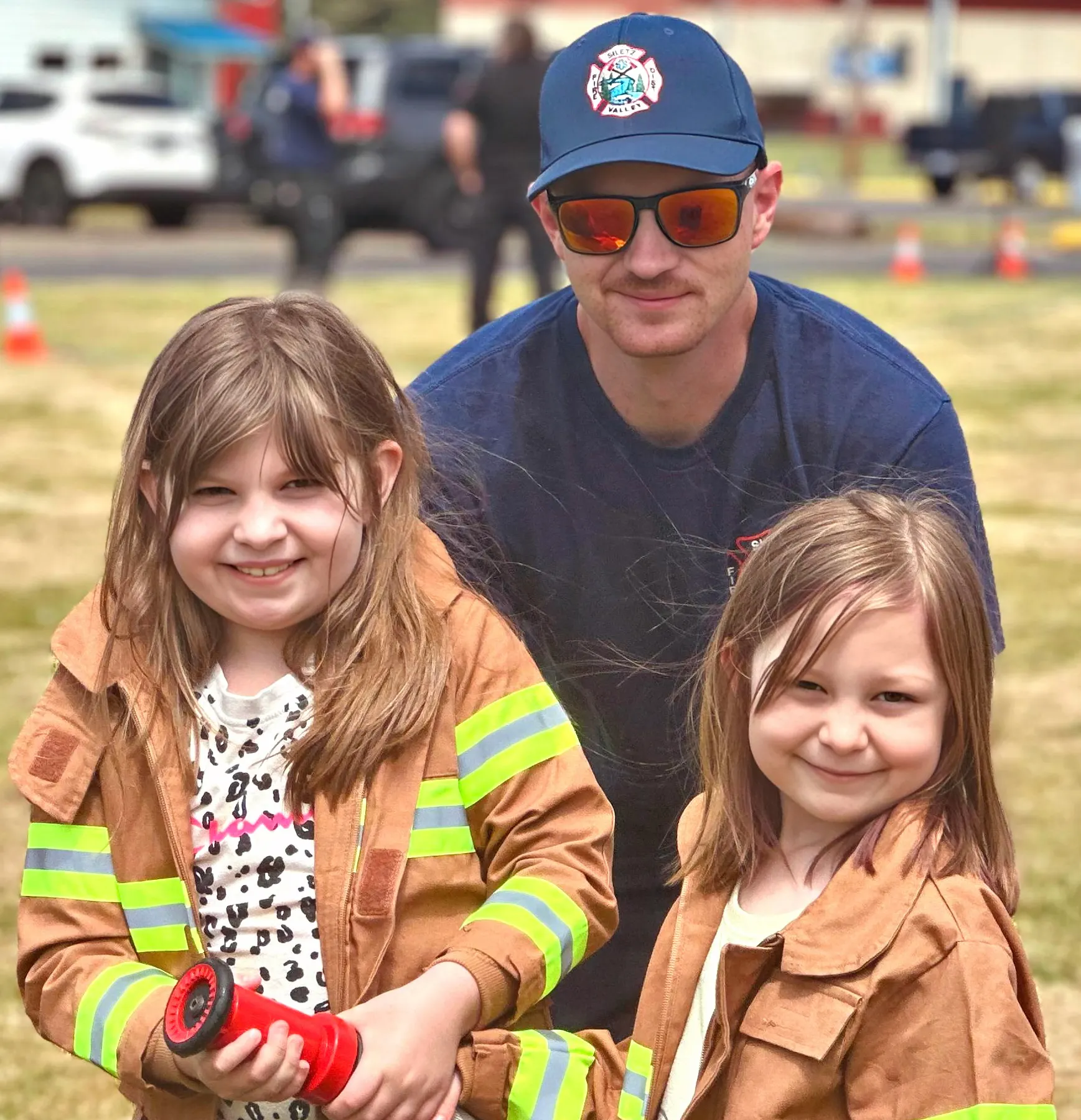 Two kids in firefighter jackets with an adult holding a hose, smiling outdoors with emergency vehicles in the background.
