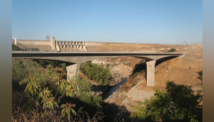 A bridge over a ravine with a dam in the background, surrounded by arid landscape.