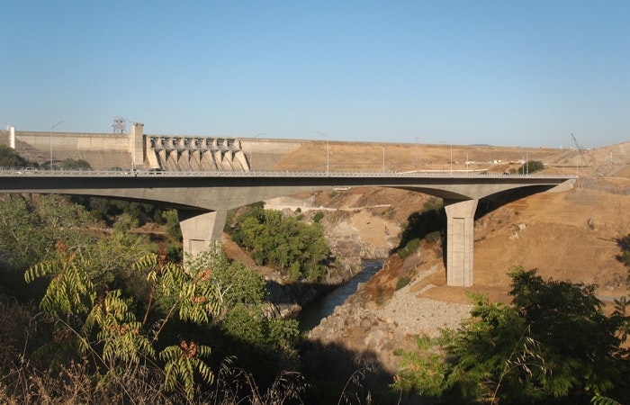 A bridge over a ravine with a dam in the background, surrounded by arid landscape.