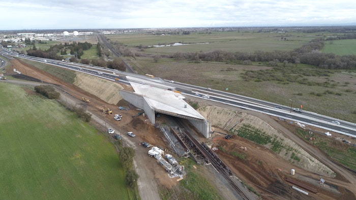 Aerial view of a highway overpass construction site with equipment and vehicles, surrounded by open fields.