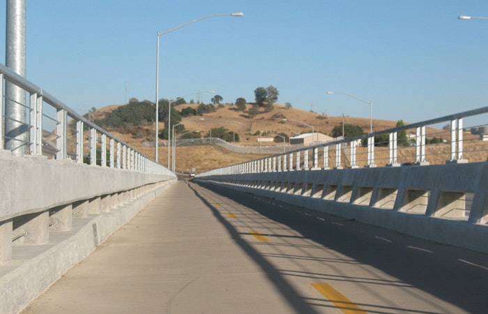 A pedestrian walkway alongside a bridge with guardrails under a clear sky, leading towards hills.