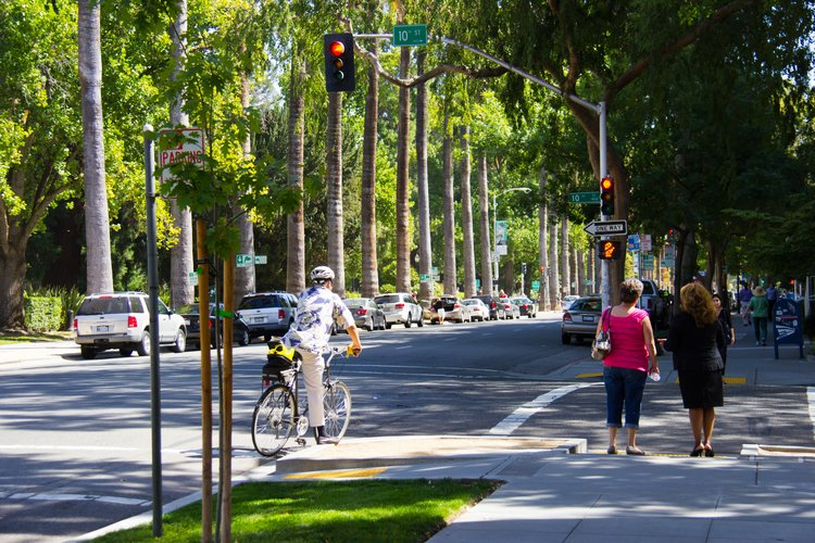 A cyclist waiting at a red light and pedestrians on a leafy street.