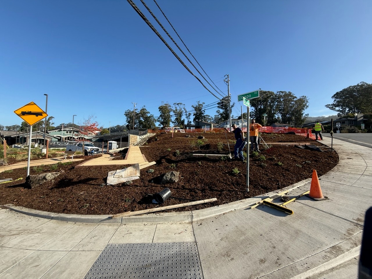 A landscaped street corner under construction with workers and orange cones, featuring a warning sign and a street sign under a clear blue sky.