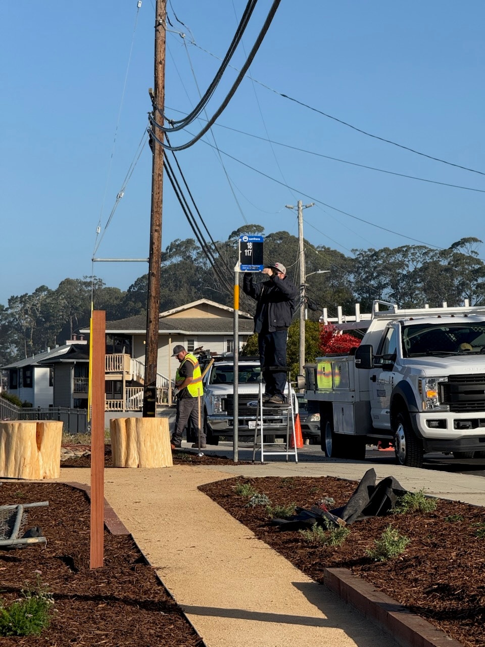 Two workers by a road: one on a ladder adjusting a sign, and another beside a truck.
