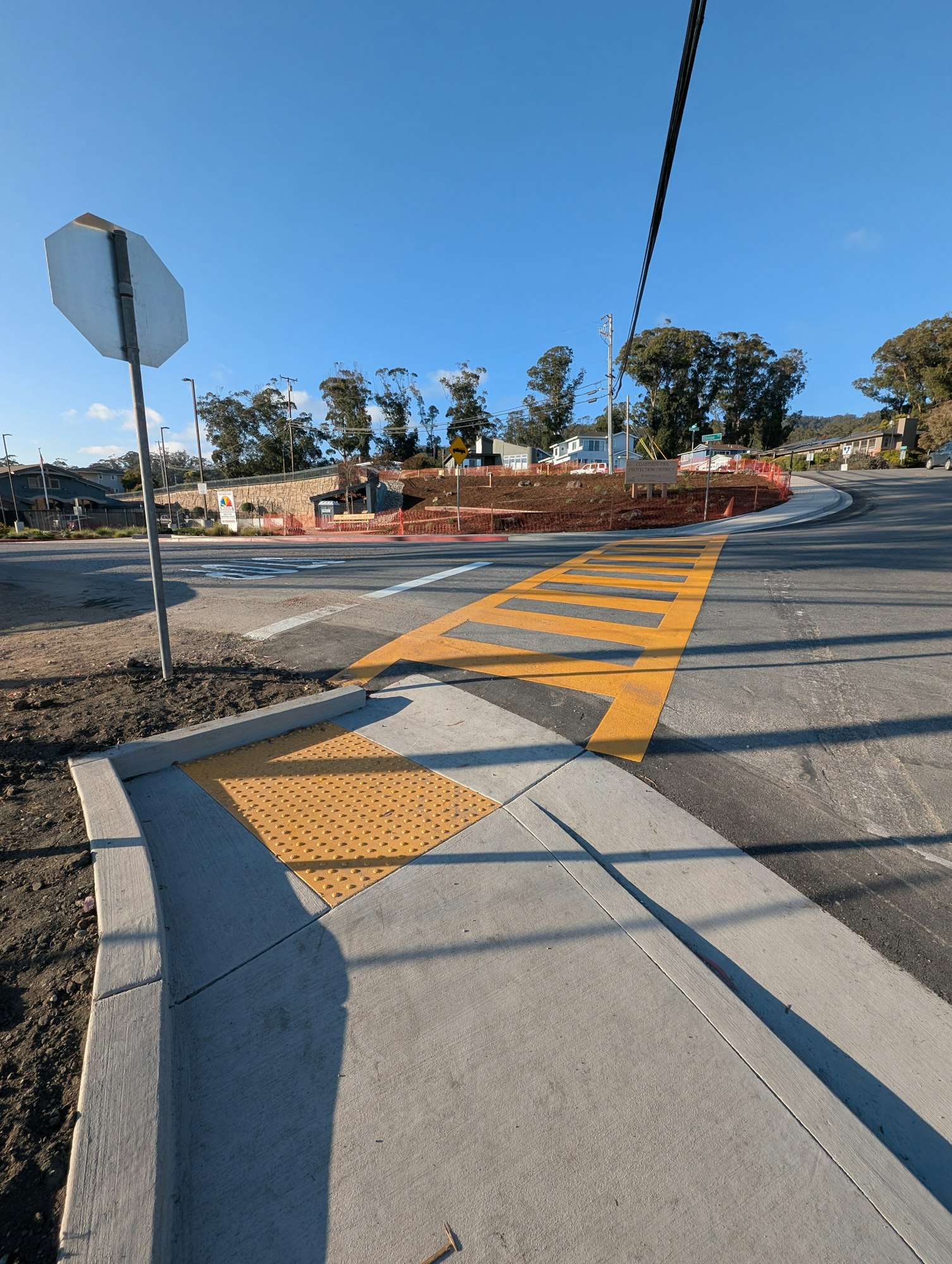 A street intersection with crosswalk, stop sign, and tactile paving for accessibility. Clear sky and trees in the background.