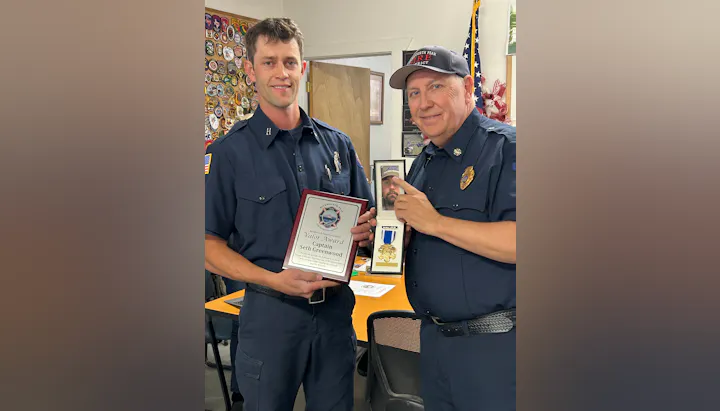 Two firefighters are celebrating an award presentation in an office, holding plaques and trophies with a backdrop of badges.