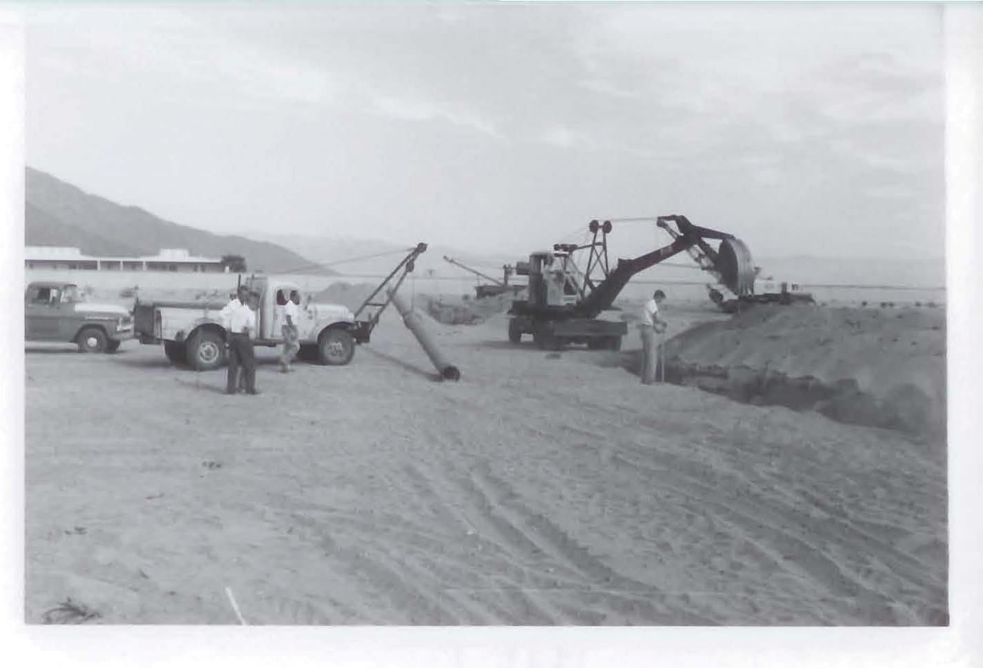 A black-and-white image showing construction work in a sandy area with machinery and workers present.