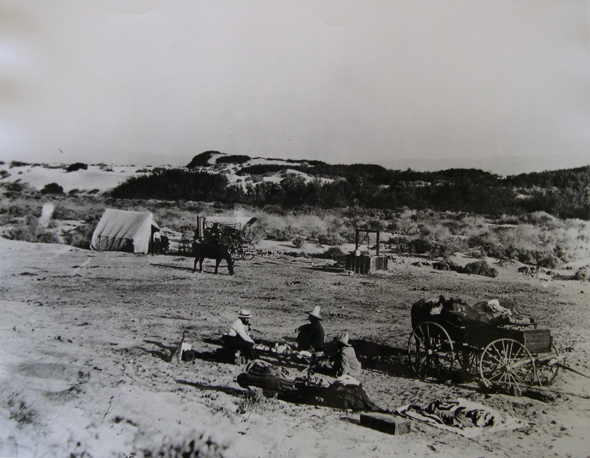 A black-and-white photo showing a dusty landscape with a tent, horse-drawn cart, and people resting.