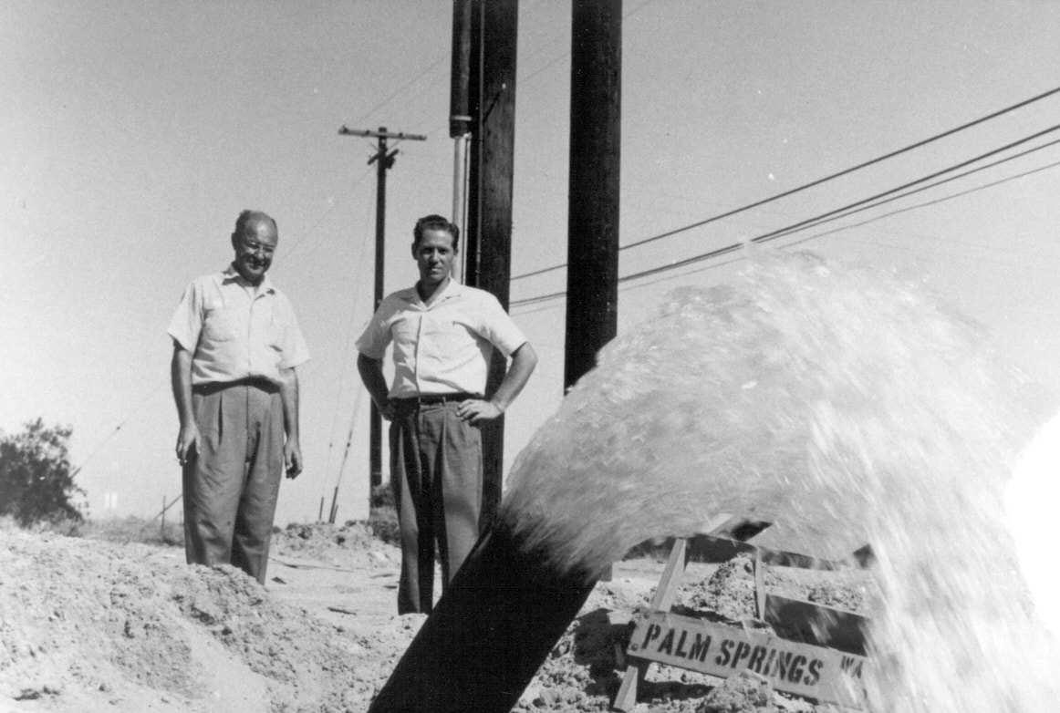 Two men stand by a pipe spewing water, with a sign for Palm Springs nearby, set against a clear sky and power lines.