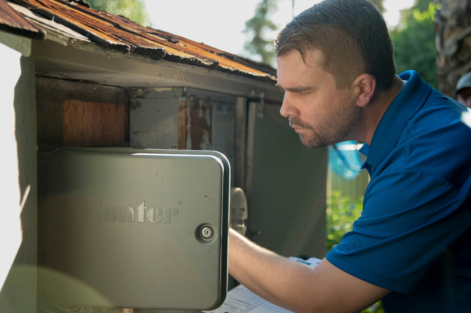 A man in a blue shirt is inspecting a Hunter irrigation control box near a house. Sunlight highlights the scene.
